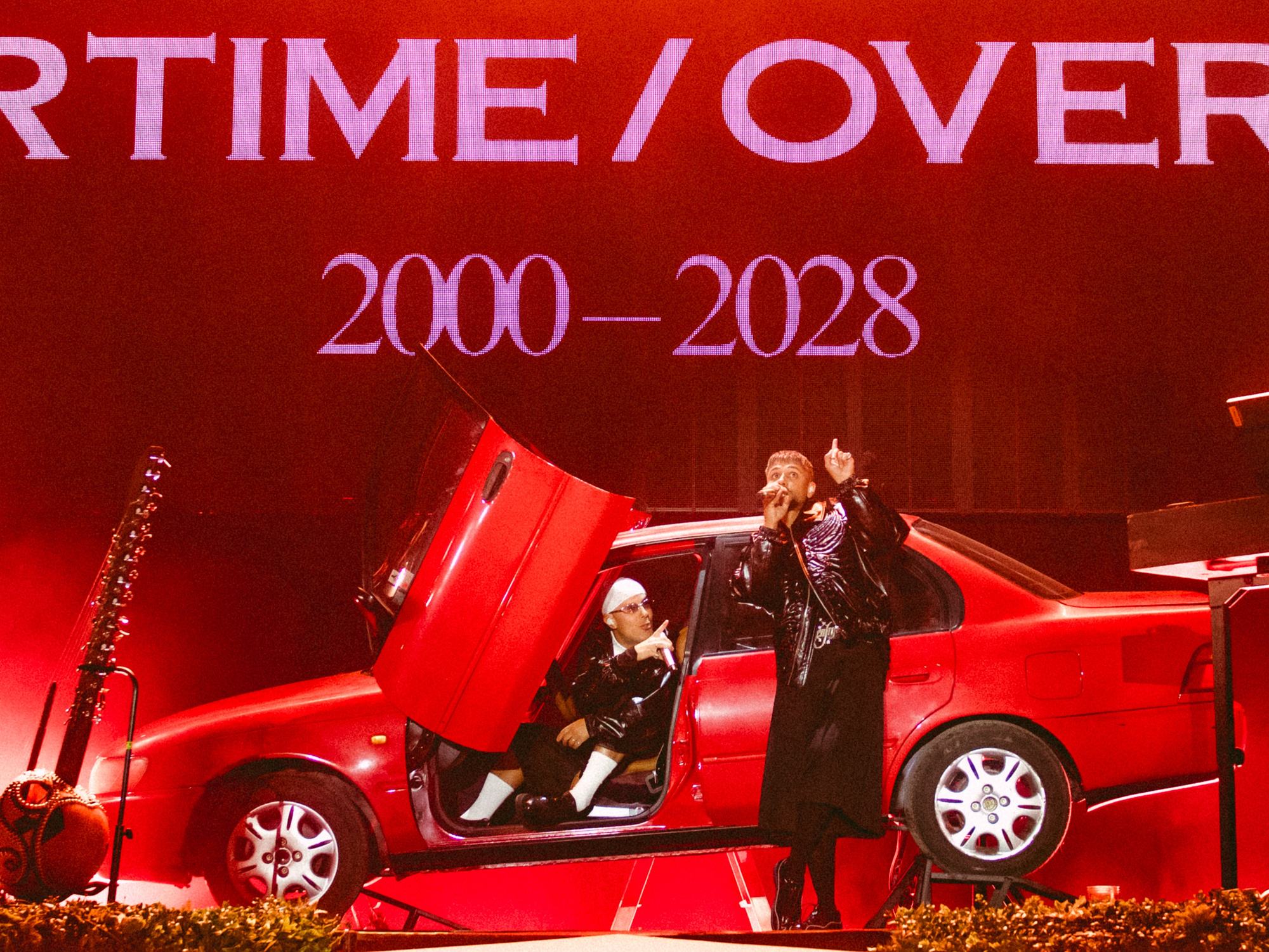 Karpe på scenen under Parkenfestivalen i Bodø i august 2026, ved en rød bil foran en rød skjerm med turnénavnet og årstallene 2000 til 2028