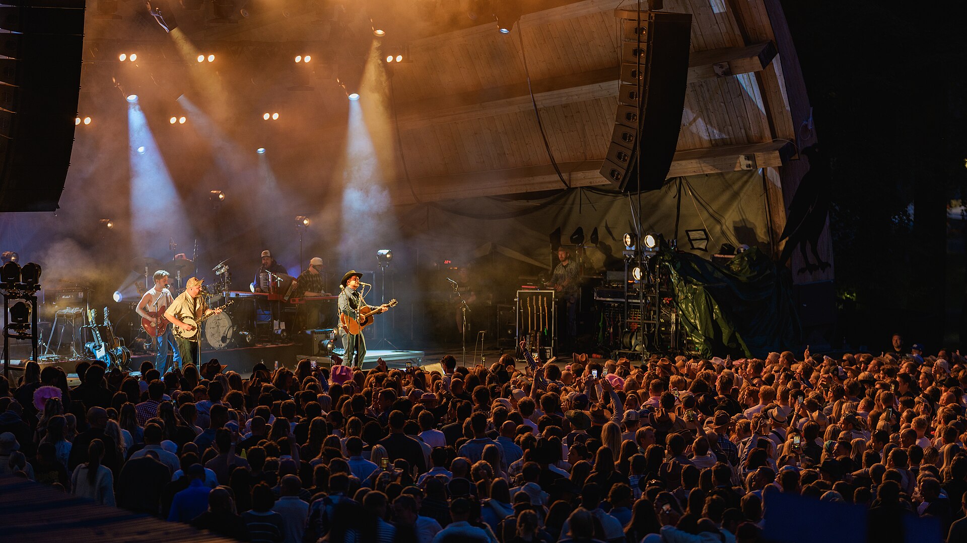 Tobias Sten med hatt og kassegitar sammen med bandet sitt på utescenen under Ravnedalen Live i Kristiansand i 2026, med fullt publikum foran scenen