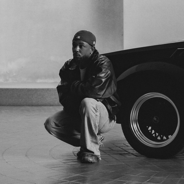 Kendrick Lamar på huk ved siden av en svart bil, i svart-hvitt