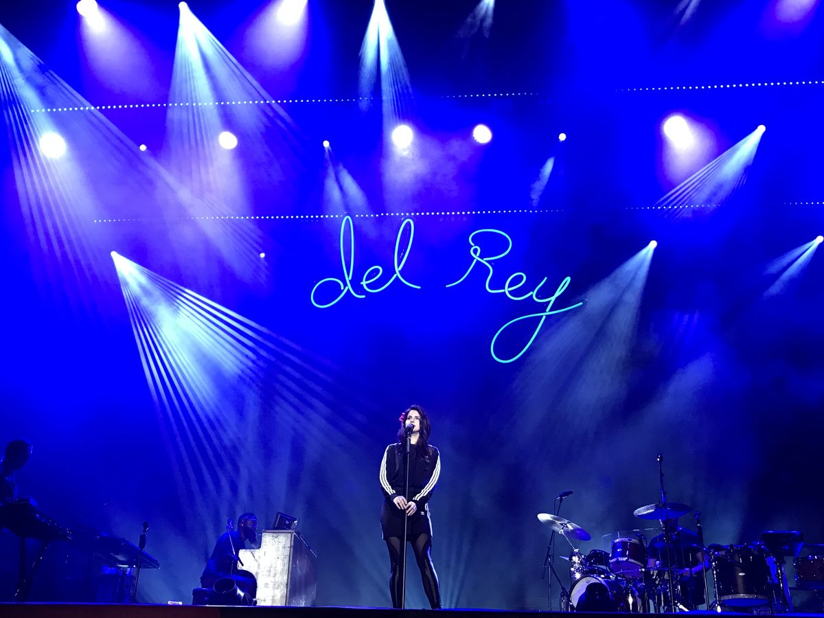 Lana Del Rey alene foran mikrofonen under blått scenelys og neonskriften «del Rey» på Flow-festivalen i Helsingfors i 2017, med band på begge sider