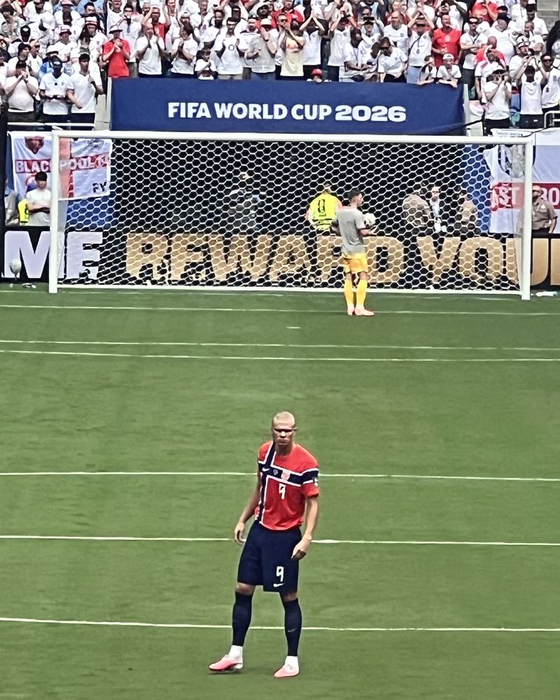 Erling Braut Haaland i rød Norge-drakt med nummer 9 står på gresset. Bak målet sitter engelske supportere i hvitt under et banner med teksten FIFA World Cup 2026.