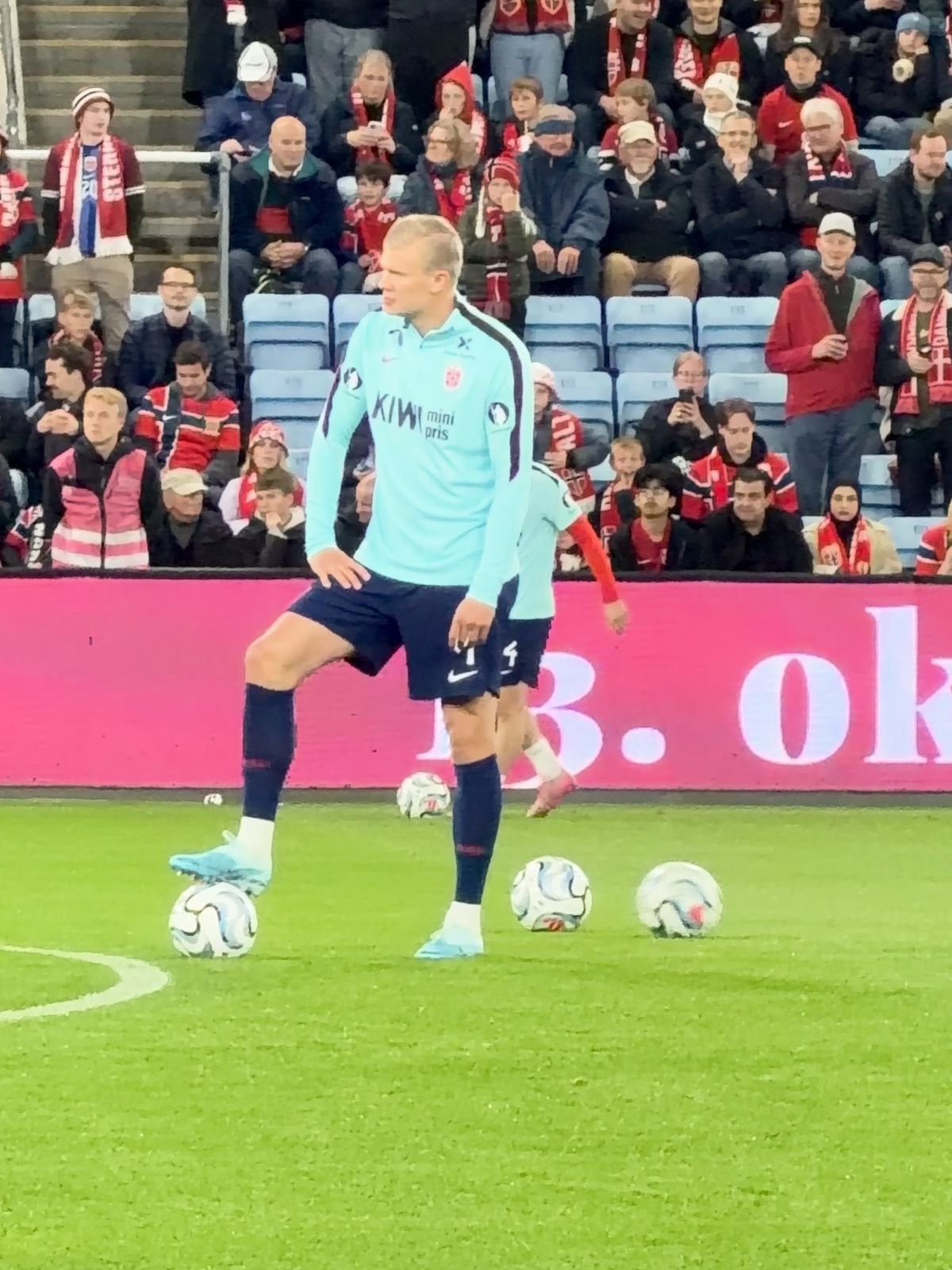Erling Braut Haaland i lyseblå treningsgenser med én fot på en ball under oppvarmingen, foran norske supportere med røde og hvite skjerf på tribunen