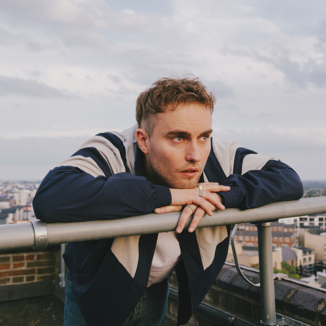 Sam Fender i mørkeblå og beige jakke lener seg på et rekkverk på et hustak