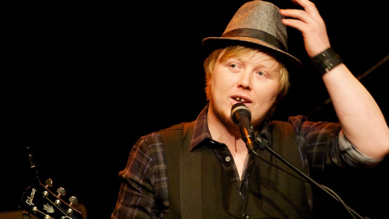 Kurt Nilsen i hatt og rutete skjorte synger i mikrofonen på en mørk scene, med en gitarhals i forgrunnen