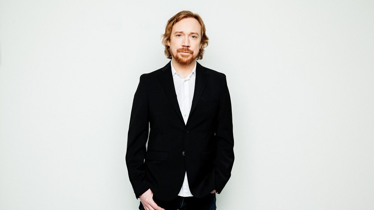 Lars Winnerbäck i svart dress og hvit skjorte foran en lys vegg, portrett fra 2016