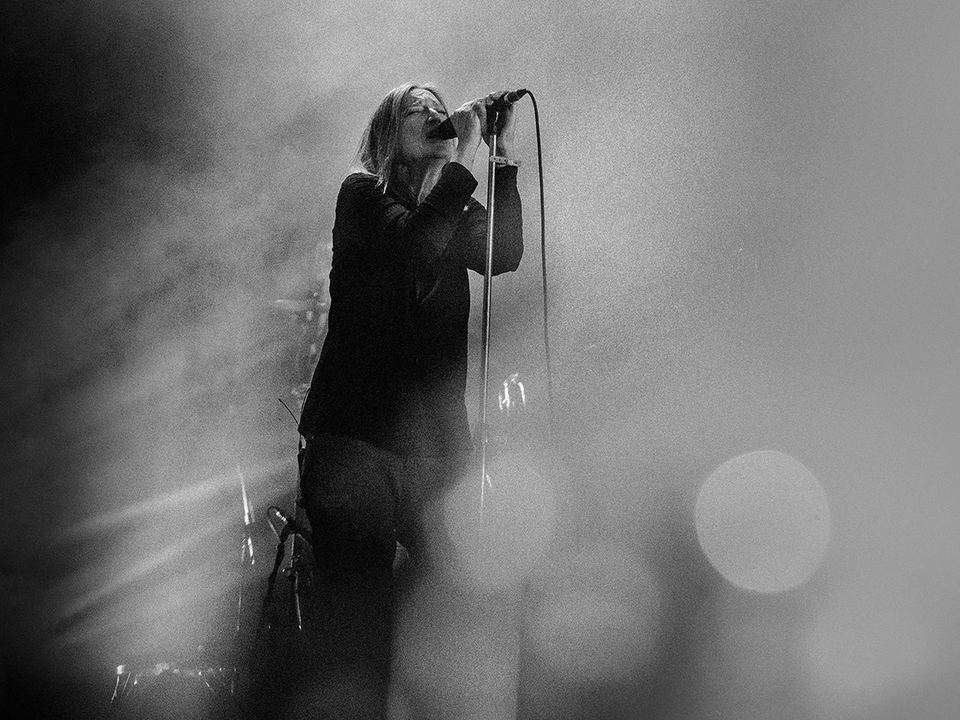 Svart-hvitt pressebilde av Beth Gibbons som synger i en mikrofon på scenen, omgitt av røyk og uskarpe lyspunkter