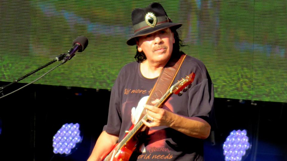 Carlos Santana med rød elgitar og svart hatt på scenen i London i 2018, foran en stor LED-skjerm
