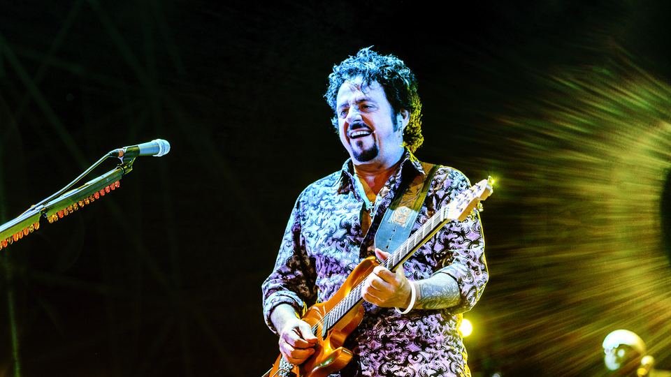 Toto-gitaristen Steve Lukather smiler og spiller en oransje gitar på scenen i Italia, i lilla og grønt scenelys